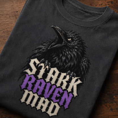 A folded black t-shirt with a graphic of a black raven and the text 'STARK RAVEN MAD' in cream and purple gothic lettering, displayed on a dark wooden surface.
