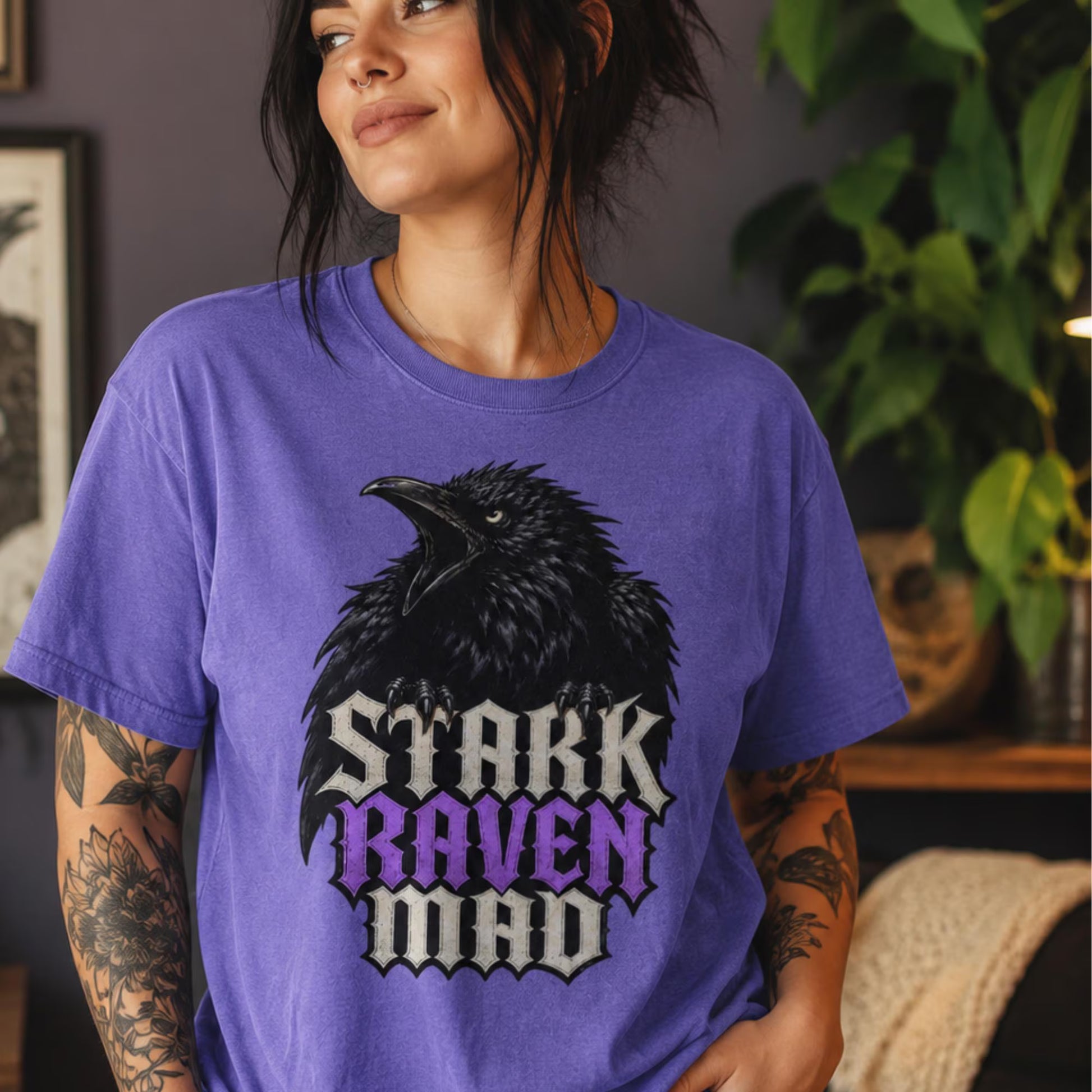 A woman wearing a purple graphic t-shirt with a black raven and the text 'STARK RAVEN MAD' in white and purple, set against a cozy indoor background with a plant.