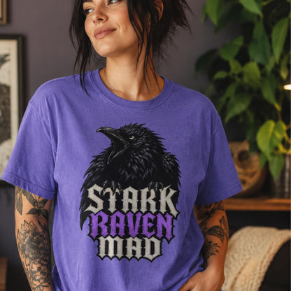 A woman wearing a purple graphic t-shirt with a black raven and the text 'STARK RAVEN MAD' in white and purple, set against a cozy indoor background with a plant.