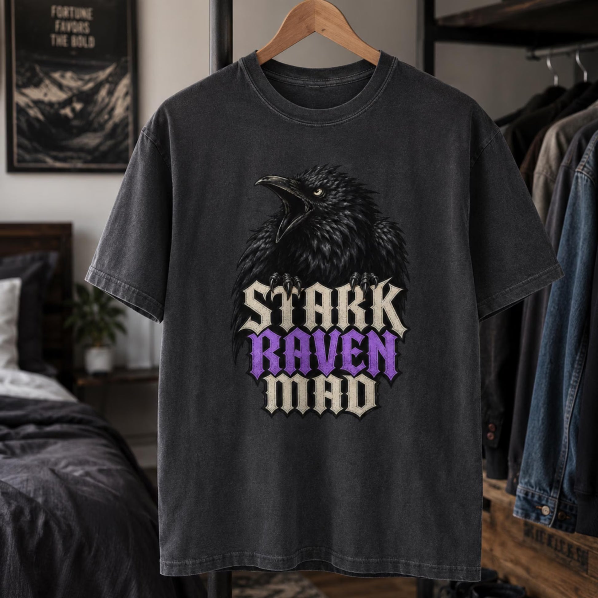 A dark gray washed graphic t-shirt hanging on a wooden hanger in a bedroom setting. The shirt features a large black raven graphic above the text 'STARK RAVEN MAD' in a stylized font, with 'RAVEN' highlighted in purple.