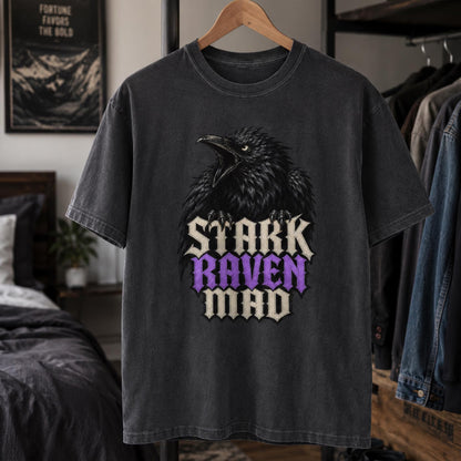 A dark gray washed graphic t-shirt hanging on a wooden hanger in a bedroom setting. The shirt features a large black raven graphic above the text 'STARK RAVEN MAD' in a stylized font, with 'RAVEN' highlighted in purple.