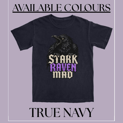 A navy blue t-shirt featuring a black raven graphic with the text 'STARK RAVEN MAD' in cream and purple. The image is framed with a light purple background and text overlays reading 'AVAILABLE COLOURS' at the top and 'TRUE NAVY' at the bottom.