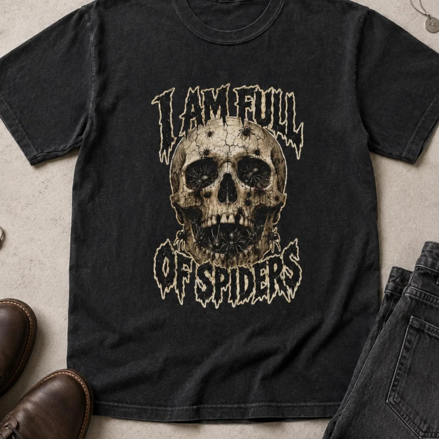 A black graphic t-shirt featuring a distressed skull design with the text 'I AM FULL OF SPIDERS' in a stylized font. The shirt is presented in a flat-lay arrangement alongside brown leather shoes, dark gray jeans, and rings on a textured concrete surface.