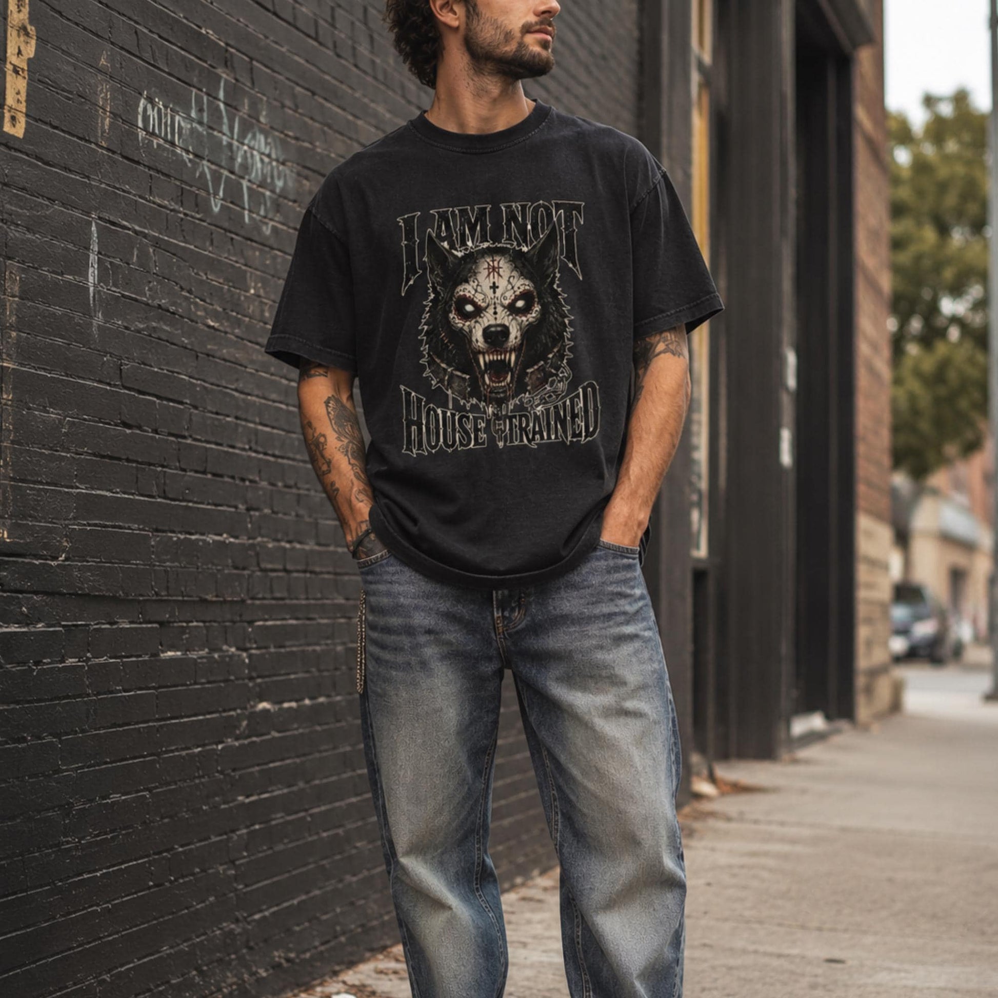 A man wearing a black graphic t-shirt with a wolf design and the words 'DOMINANT HOUSE TRAINED', paired with blue jeans, standing on a city sidewalk next to a dark brick wall.