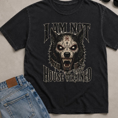 A black graphic t-shirt featuring a stylized snarling wolf with skull-like markings and the text 'I AM NOT HOUSE TRAINED' in a distressed font. The t-shirt is styled in a flat-lay arrangement with blue jeans on a neutral gray background.