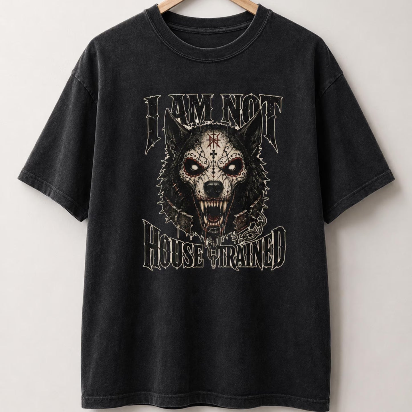 A black washed graphic t-shirt hanging on a wooden hanger against a light gray wall. The shirt features a large front graphic of a skull-faced snarling wolf with a red cross on its forehead, framed by the text 'I AM NOT HOUSE TRAINED' in a distressed gothic font.