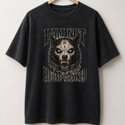 A black washed graphic t-shirt hanging on a wooden hanger against a light gray wall. The shirt features a large front graphic of a skull-faced snarling wolf with a red cross on its forehead, framed by the text 'I AM NOT HOUSE TRAINED' in a distressed gothic font.