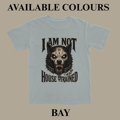 A light blue-gray graphic t-shirt displayed on a solid taupe background, featuring a black and white wolf skull design with the text 'I AM NOT HOUSE TRAINED'. The image includes text overlays at the top and bottom reading 'AVAILABLE COLOURS' and 'BAY'.