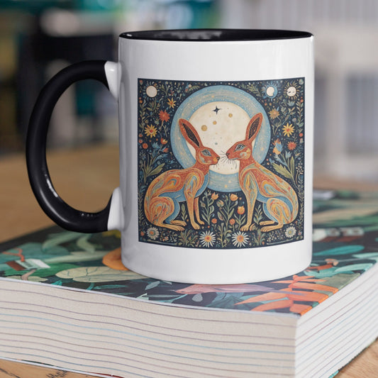 Moon gazing hares on full moon. A white 11 oz ceramic mug with a black interior and black handle. The mug features a folk art design of two hares facing each other in front of a glowing full moon, creating a whimsical and rustic aesthetic.