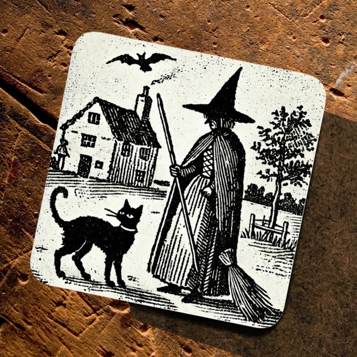 witchy gifts: woodcut of a witch with her familiar, a black cat printed on a cork backed, hardwood coaster. Witches house, home in the background with black cat wearing a collar.