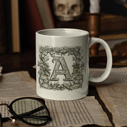 A white 11 oz mug featuring an antique-style letter  designed with intricate calligraphy and surrounded by a decorative botanical frame.