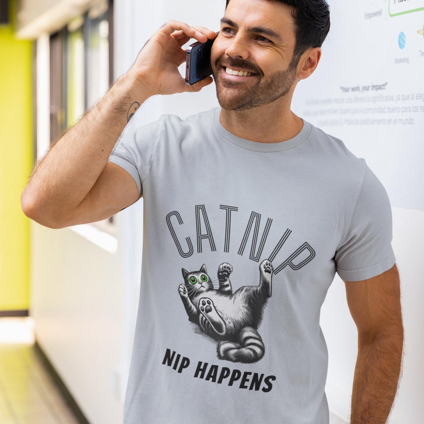 t-shirt featuring a playful design with the word CATNIP in bold, arched letters. Below, a cartoon-style cat with bright green eyes is lying on its back, looking surprised and playful. The phrase NIP HAPPENS is written underneath in bold letters.
