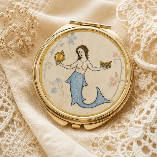 A round gold-toned compact mirror with a vintage-style illustration of a mermaid on the lid, placed on cream lace fabric with dried flowers.
