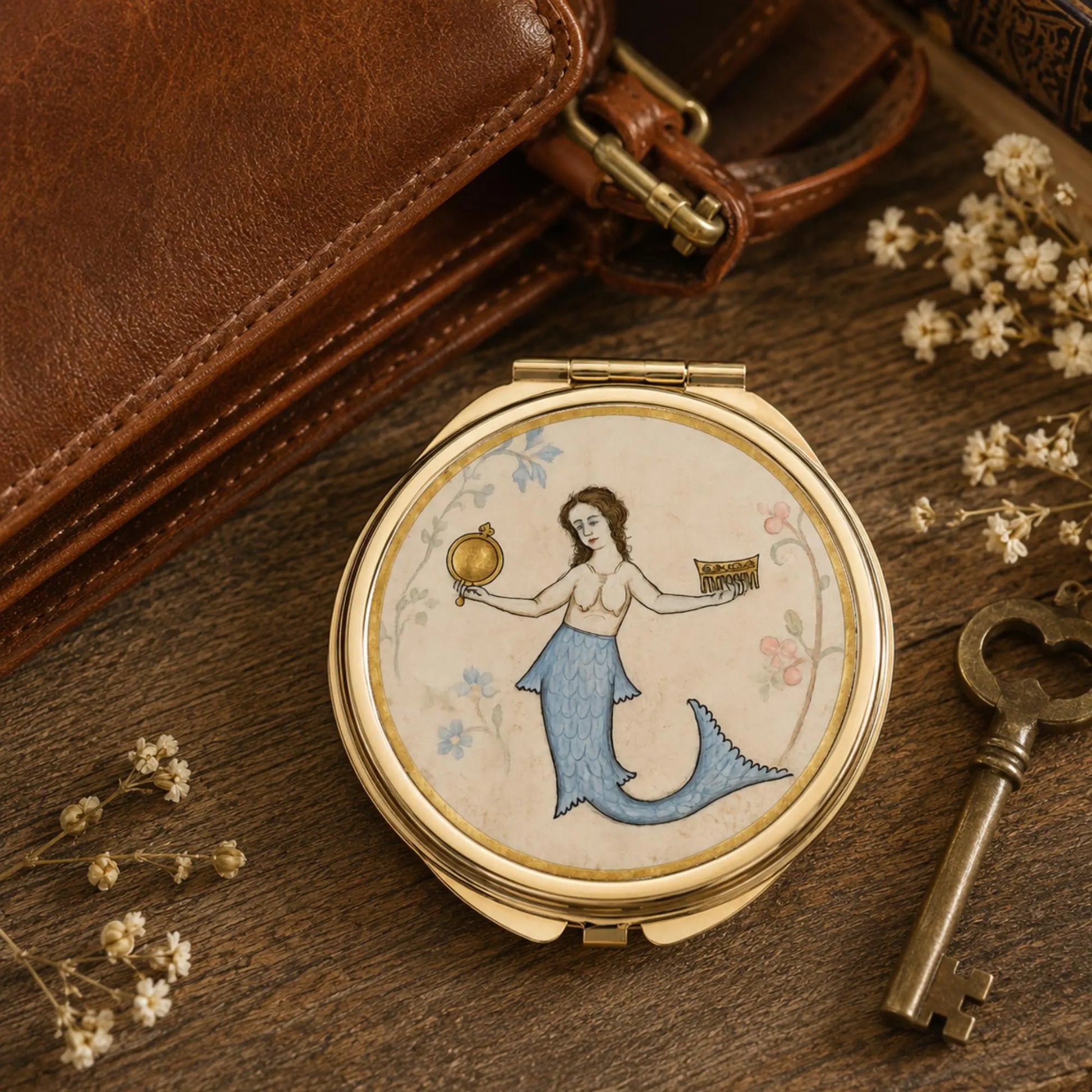 A round gold-toned compact mirror with a vintage-style illustration of a mermaid on the lid, styled on a wooden surface with a brown leather bag, a brass key, and small white flowers.