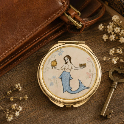 A round gold-toned compact mirror with a vintage-style illustration of a mermaid on the lid, styled on a wooden surface with a brown leather bag, a brass key, and small white flowers.