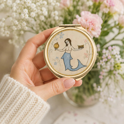 A hand wearing a cream knit sweater holds a round gold-framed compact mirror featuring a vintage-style illustration of a mermaid with a blue tail on a cream background. The scene is softly blurred with pink flowers and white baby's breath in the background.