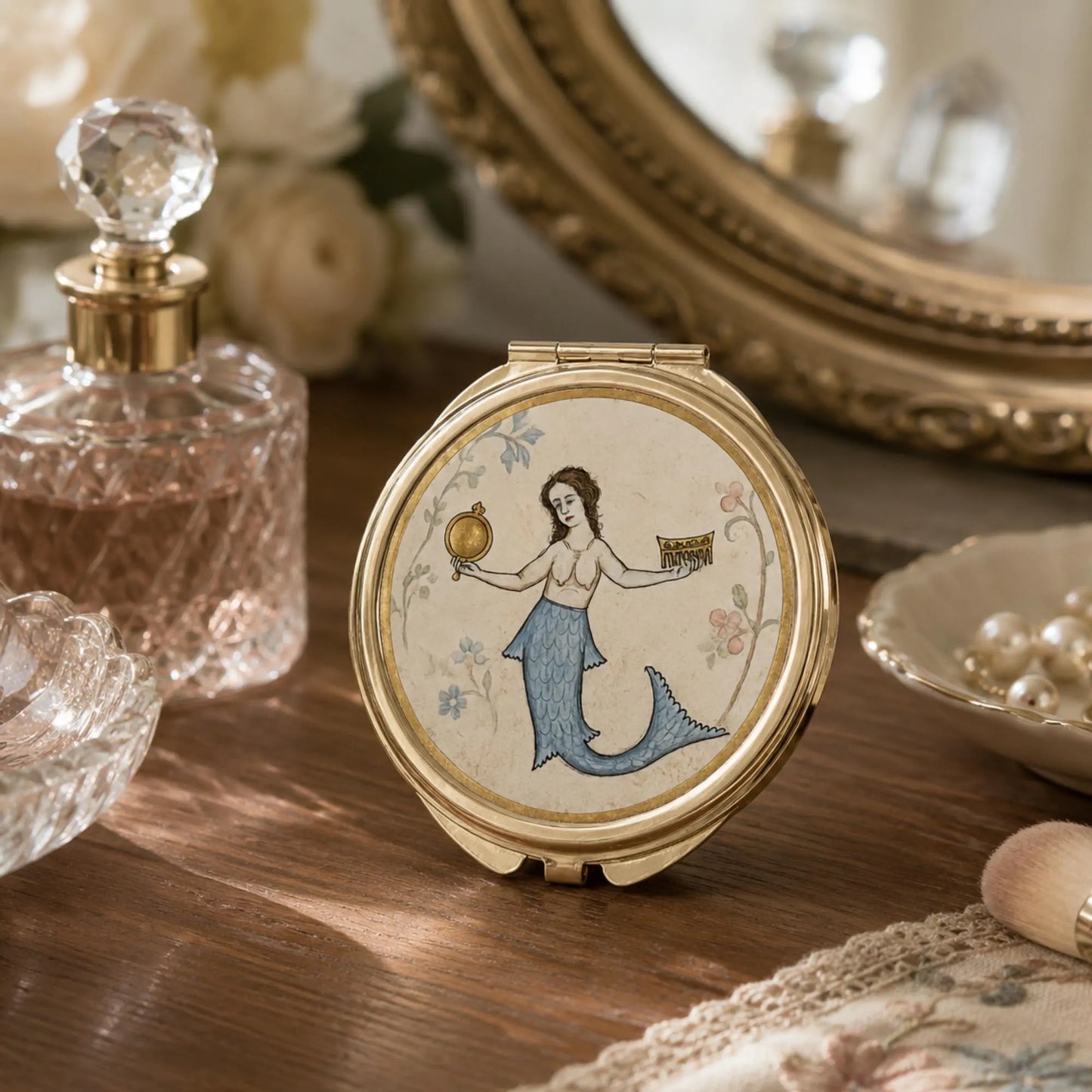 A round gold-tone folding compact mirror with a painted mermaid illustration on the lid, displayed on a wooden surface with vintage-style props including a perfume bottle, mirror, and pearls.