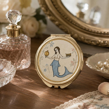 A round gold-tone folding compact mirror with a painted mermaid illustration on the lid, displayed on a wooden surface with vintage-style props including a perfume bottle, mirror, and pearls.