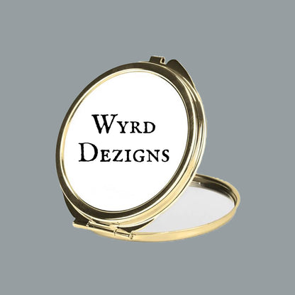 A round gold-framed compact mirror shown open against a solid gray background, with the words 'Wyrd Dezigns' printed in black on the white interior lid.