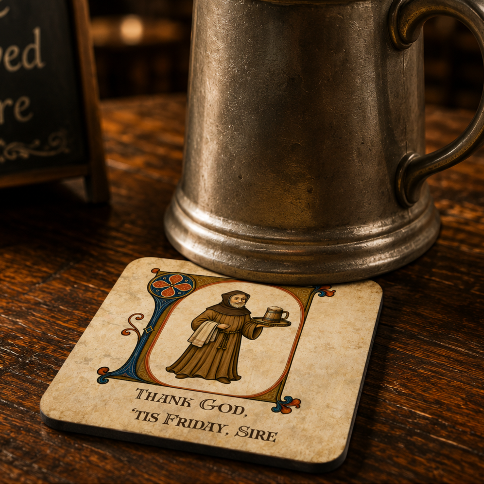 A square coaster with a medieval-style illustration of a Franciscan holding a cup, with the text 'THANK GOD, TIS FRIDAY, SIRE,' placed on a wooden table next to an antique metal mug.