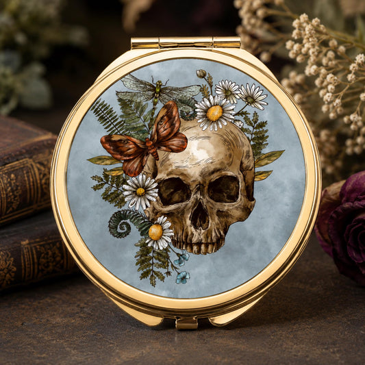 Round gold compact mirror with a light blue lid featuring a vintage-style illustration of a human skull surrounded by white daisies, green ferns, a brown butterfly, and a dragonfly, displayed on a rustic wooden surface with dried flowers and books in the background.