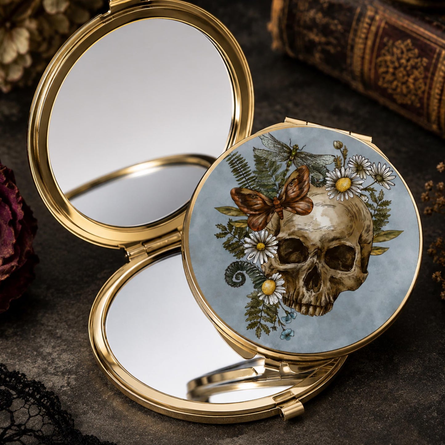 An open round gold-tone compact mirror with a decorative lid featuring a skull, daisies, ferns, and a butterfly on a pale blue background, styled on a dark textured surface with vintage books and dried flowers.