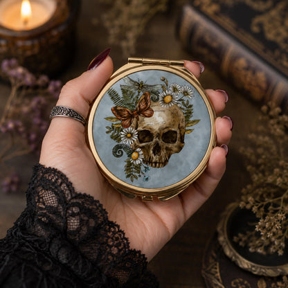 A hand with dark red nail polish holds a round gold compact mirror featuring a detailed illustration of a human skull surrounded by white daisies, a brown butterfly, and green leaves on a pale blue background. The scene is styled with a black lace sleeve, a silver ring, a lit candle, and vintage books in the background.