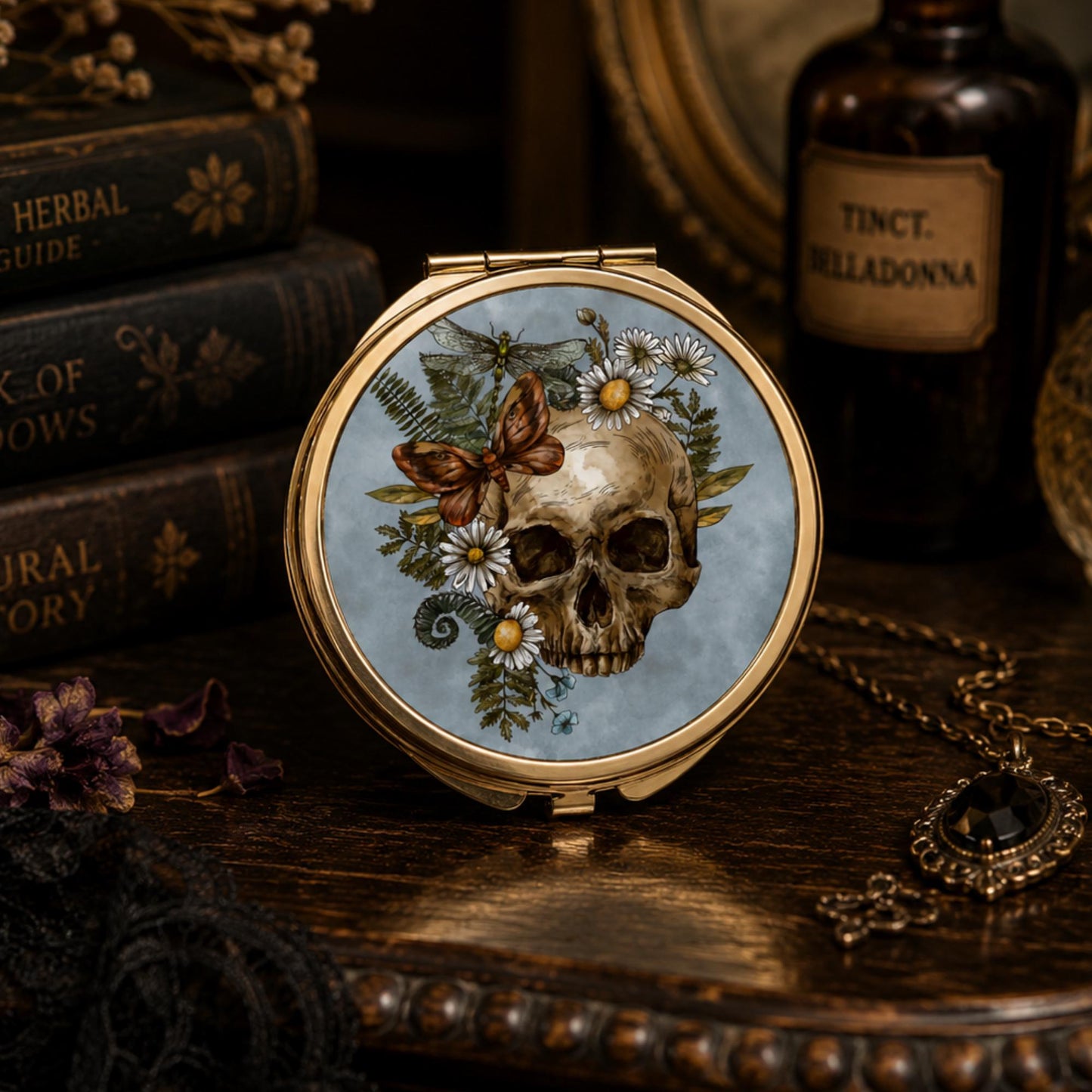 A round compact mirror with a gold-tone frame, featuring a vintage-style illustration of a human skull surrounded by white daisies, green leaves, and a brown butterfly on a light blue background. The compact is styled on a dark wooden table with antique books, a bottle of belladonna, and a necklace in the background.