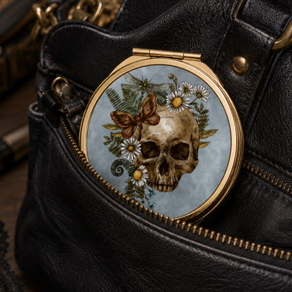A round compact mirror with a gold-tone frame and a detailed illustration of a skull, daisies, ferns, and a butterfly on a light blue background, shown resting inside the zippered pocket of a black leather bag.
