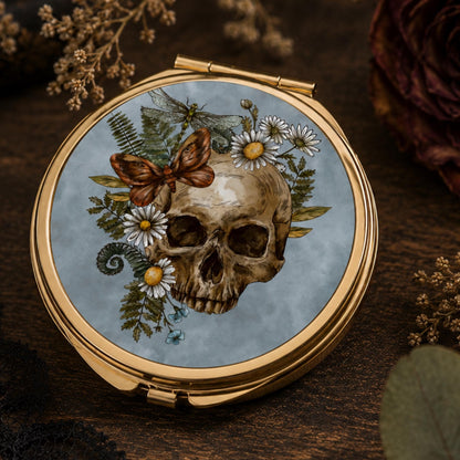 A round compact mirror with a gold-tone frame, featuring a printed illustration of a human skull surrounded by white daisies, green ferns, a brown butterfly, and a dragonfly on a pale blue background, displayed on a dark wooden surface.
