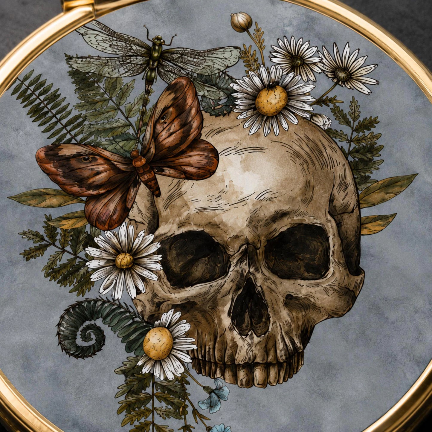 An ornate circular gold-framed artwork featuring a detailed illustration of a human skull surrounded by white daisies, green ferns, a brown butterfly, and a dragonfly on a textured gray background.