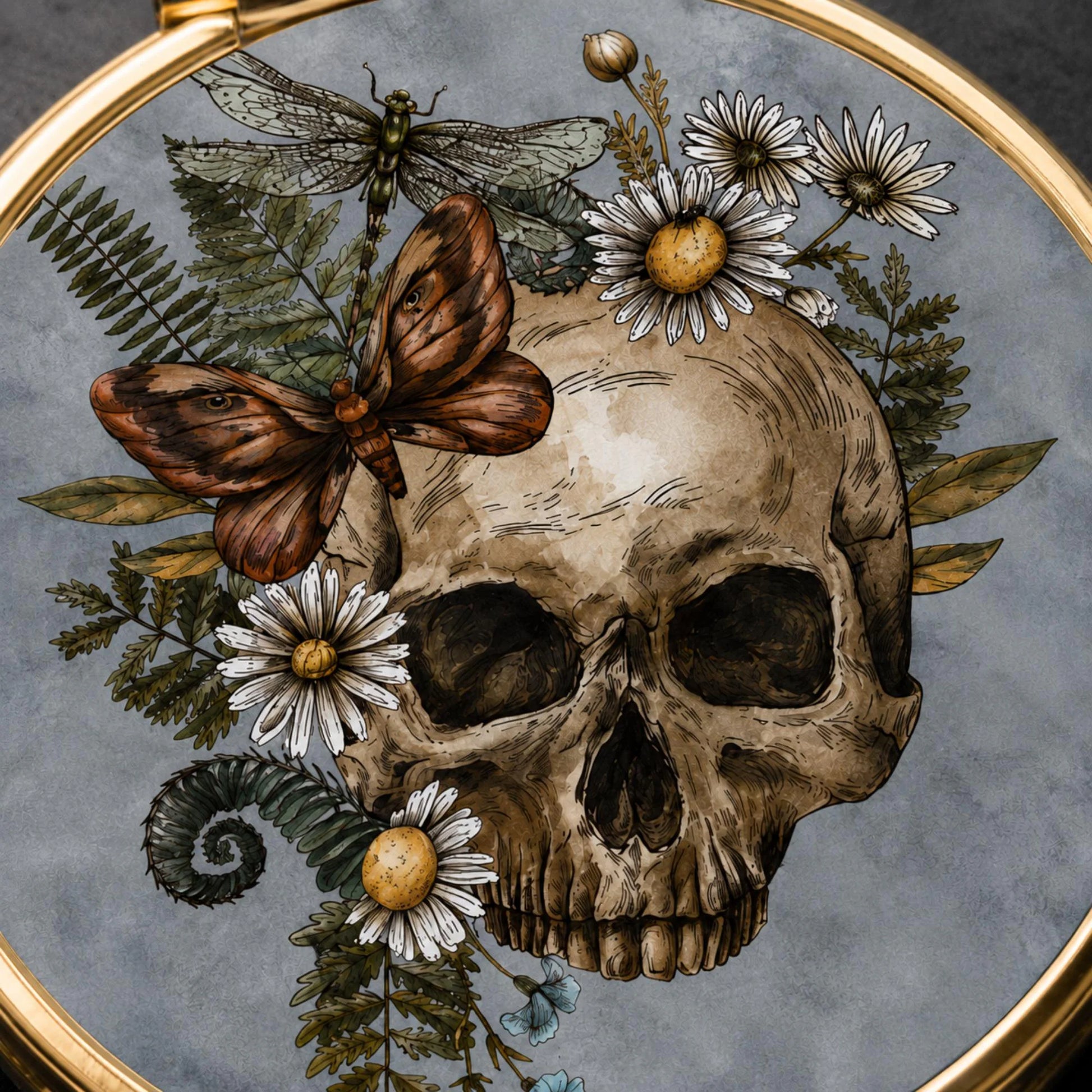 An ornate circular gold-framed artwork featuring a detailed illustration of a human skull surrounded by white daisies, green ferns, a brown butterfly, and a dragonfly on a textured gray background.